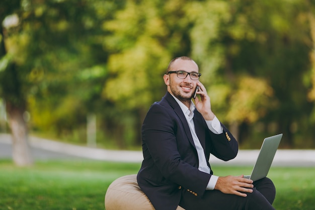 Young businessman in white shirt, classic suit, glasses. Man sit on soft pouf, talk on phone, work on laptop pc computer in city park on green lawn outdoors on nature. Mobile Office, business concept.