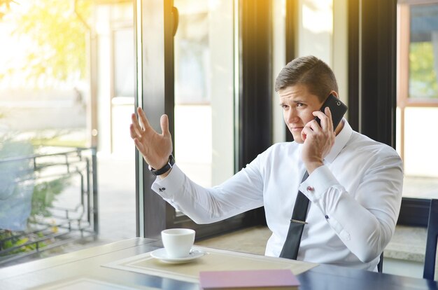 Young businessman in a white shirt and tie is sitting in a cafe in the morning and talking on the phone A cup of coffee is on the table