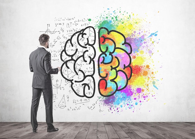 young businessman with a beard wearing a gray suit is standing and holding papers. he is looking forward. a concrete wall with a colorful brain sketch