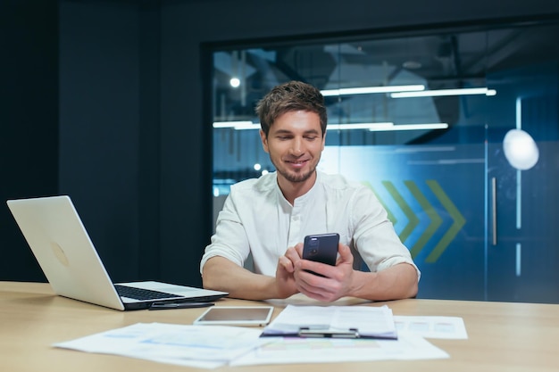 Молодой бизнесмен с бородой работает в современном офисе с ноутбуком, смотрит на экран телефона, улыбается, читает хорошие новости
