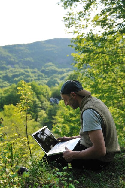 young businessman with hat work on thin laptop outdoor in nature with beautiful waterfalls in background representing freedom and wireless technology concept