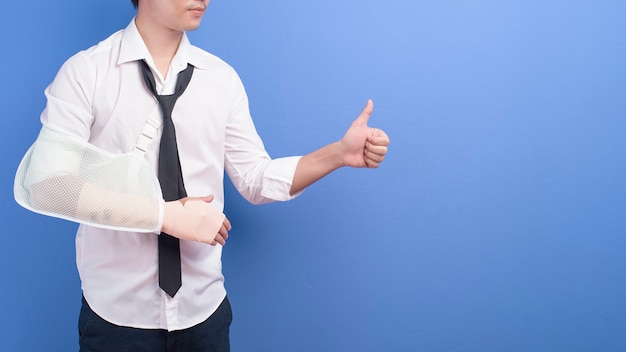 A young businessman with an injured arm in a sling over blue wall, insurance and healthcare concept