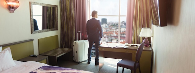 ホテルの部屋で荷物を持つ青年実業家。窓を見て、出張や休暇に到着し、スペースをコピーするスーツを着た成功した男