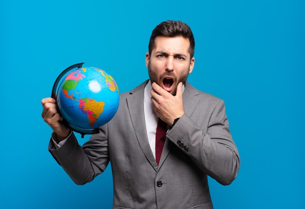 Young businessman with mouth and eyes wide open and hand on chin, feeling unpleasantly shocked, saying what or wow holding a world globe map