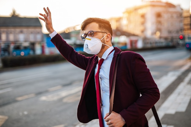 Young businessman with protective mask standing alone on empty street and waiting for bus or taxi. Virus pandemic or pollution concept.