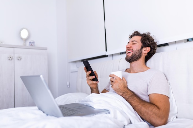 Young businessman working happy and relaxed from bed with laptop computer using mobile phone in workaholic concept , Working from home in the morrning and drinking coffe