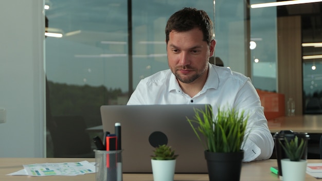 Young businessman working on laptop at desk in modern open plan start up office being joined by female colleague for informal meeting