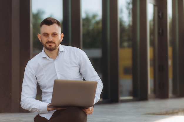 Молодой бизнесмен работает в Интернете с ноутбуком, сидя на скамейке за пределами своего офисного здания