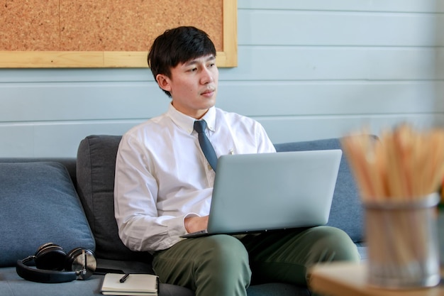 Young businessman working with laptop earphone and paper note on resting couch of office lobby and close eyes to think solution about marketing strategy and planning network to solve difficult task