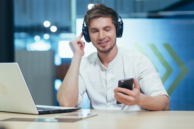 Young businessman works in the office on a laptop listens to music in big headphones uses the application to listen to online books and podcasts uses the phone