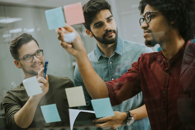 Giovani uomini d'affari che discutono davanti alla parete di vetro usando le note e gli adesivi di post-it