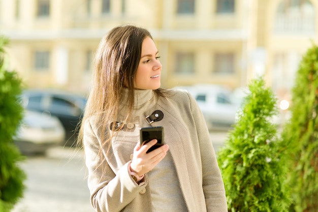 Young businesswoman in autumn city in coat with smartphone on street. City portrait of successful fashionable beautiful female