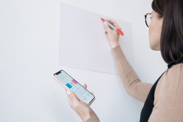 Young businesswoman or coach holding smartphone with flow chart while going to rewrite it on whiteboard at seminar