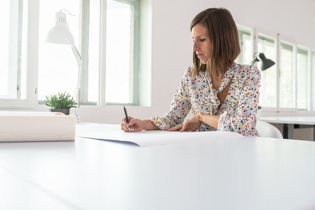 Young businesswoman or designer sitting at her office desk working on a project drawing with pencil on a large piece of paper.