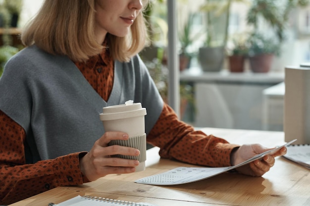 Young businesswoman drinking coffee from disposable cup while sitting at table and reading document