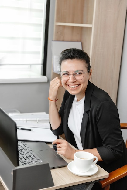 A young businesswoman employee or manager uses a computer looks at a customer or employee and smiles Hybrid work empty office