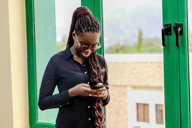 Молодая бизнесменка, занятая своим телефоном, стоя у окна.