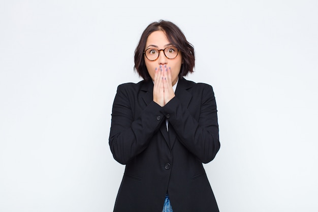 Young businesswoman feeling worried, upset and scared, covering mouth with hands, looking anxious and having messed up against white wall