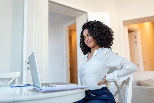 Young businesswoman having back pain while sitting at office\
desk businesswoman holding her back while working on laptop at\
office desk