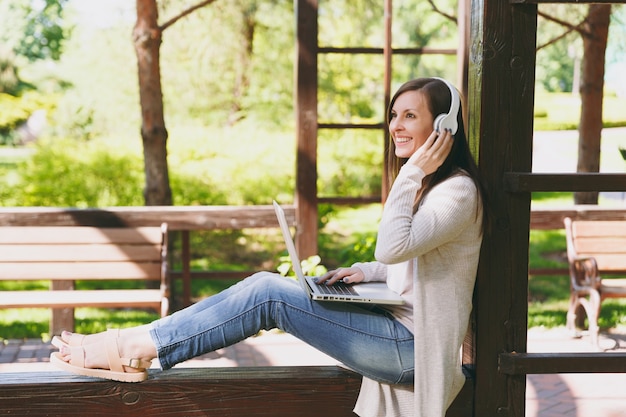 Young businesswoman in light casual clothes. Woman working on modern laptop pc computer, listen music in headphones on head in street outdoors. Mobile Office. Freelance business concept. Side view.