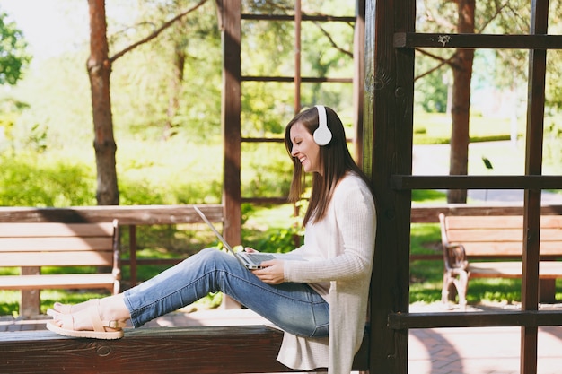 Young businesswoman in light casual clothes. Woman working on modern laptop pc computer, listen music in headphones on head in street outdoors. Mobile Office. Freelance business concept. Side view.