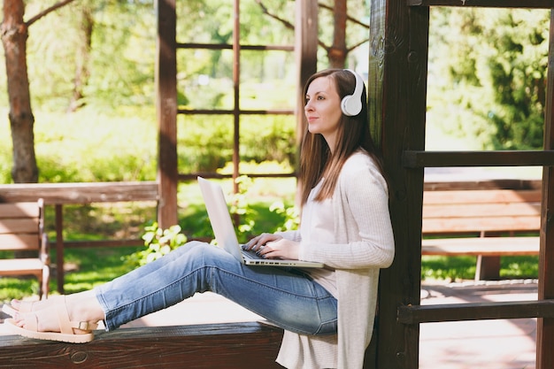 Young businesswoman in light casual clothes. Woman working on modern laptop pc computer, listen music in headphones on head in street outdoors. Mobile Office. Freelance business concept. Side view.