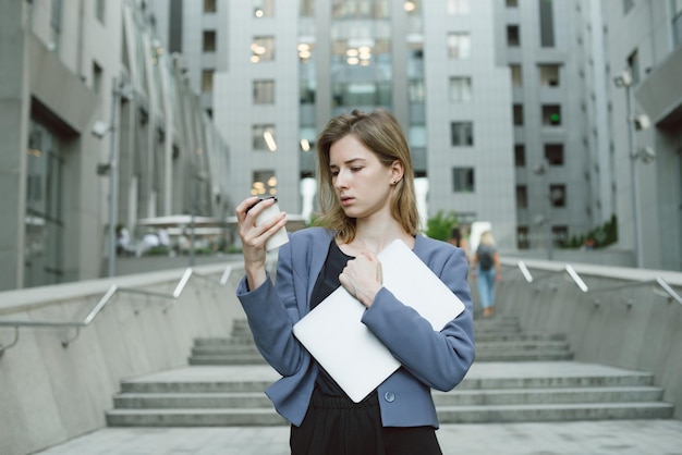 Young businesswoman looking at cup of coffee holding her laptop near business center