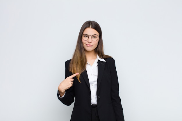 Young businesswoman looking proud, confident and happy, smiling and pointing to self or making number one sign against white wall