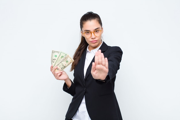 Young businesswoman looking serious, stern, displeased and angry showing open palm making stop gesture with banknotes with bills