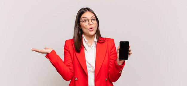 Young businesswoman looking surprised and shocked, with jaw dropped holding an object with an open hand on the side