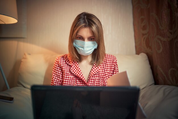 Young businesswoman in a medical protective mask works from home at the computer during self-isolation and quarantine