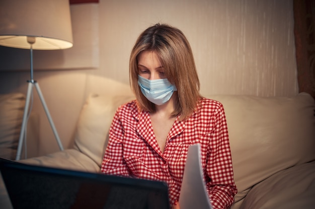 Young businesswoman in a medical protective mask works from home at the computer during self-isolation and quarantine.