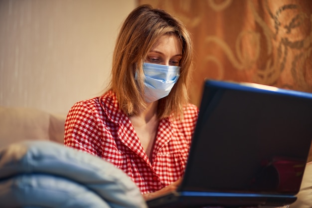 Young businesswoman in a medical protective mask works from home at the computer during self-isolation and quarantine.