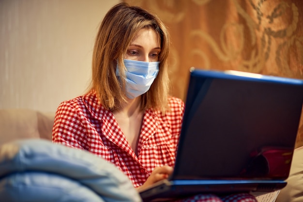 Young businesswoman in a medical protective mask works from home at the computer during self-isolation and quarantine.