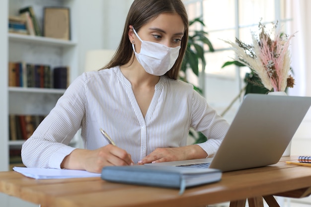 Young businesswoman in a medical protective mask works from home at the laptop during self-isolation and quarantine.