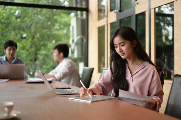 Young businesswoman at modern startup office working on tablet, blured team in meeting background.