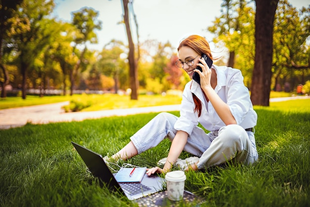 La giovane donna d'affari si siede sull'erba nel parco usa il pc portatile e il cellulare per lavorare fuori dall'ufficio