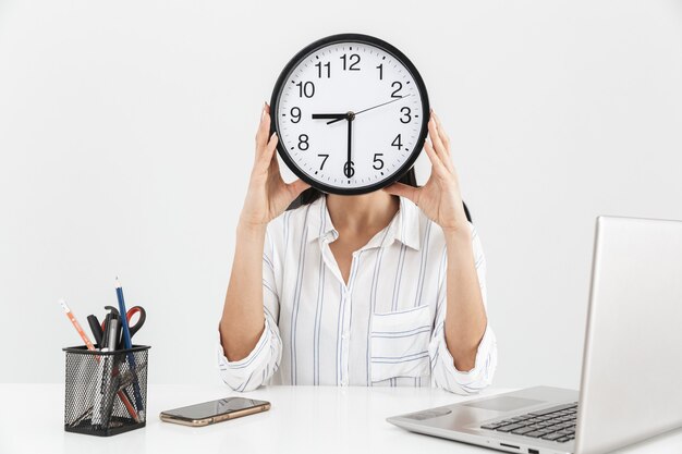 Foto giovane imprenditrice seduto in poltrona in ufficio e tenendo il grande orologio davanti al viso isolato sul muro bianco