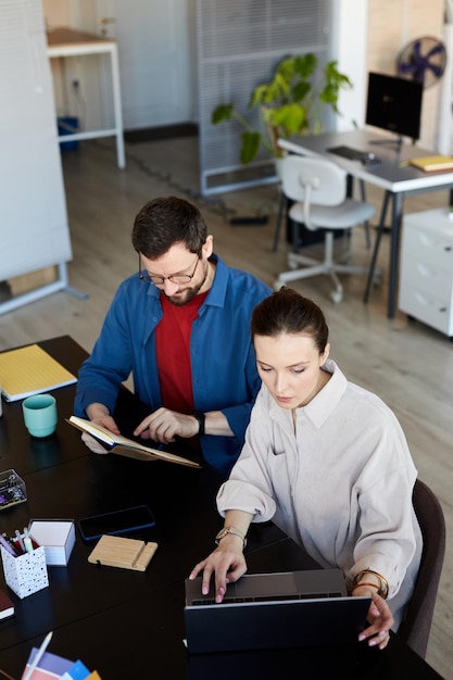 Young businesswoman sitting in front of laptop and networking while her colleague looking through no