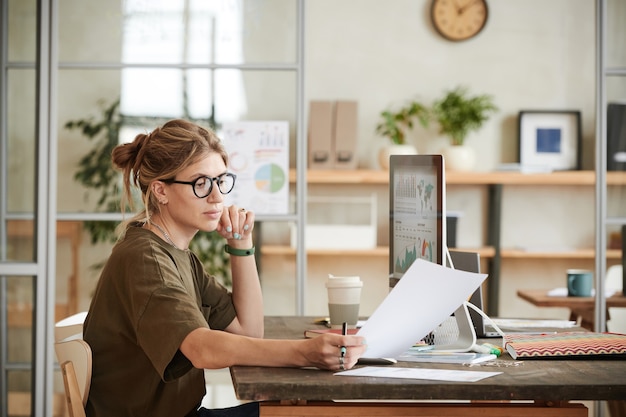 コンピューターのモニターの前で彼女の職場に座って、オフィスでドキュメントを扱う若い実業家