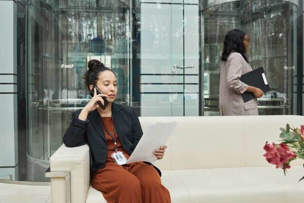 Young businesswoman sitting on sofa in the office corridor reading document and talking on mobile phone