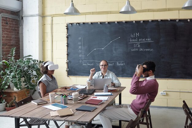 Young businesswoman sitting between two colleagues in vr headsets and looking at one of them during presentation