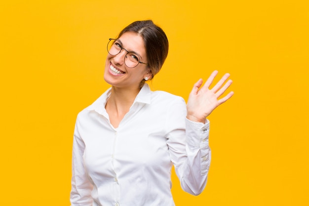 Giovane imprenditrice sorridendo felicemente e allegramente, agitando la mano, dandovi il benvenuto e salutandovi, o salutandovi contro il muro arancione