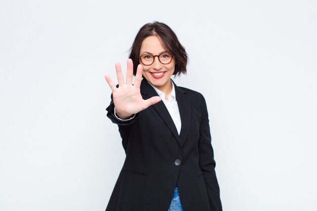 Young businesswoman smiling and looking friendly, showing number five or fifth with hand forward, counting down over white wall