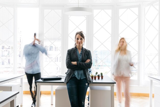 Young businesswoman standing with arms crossed in office while colleagues are moving