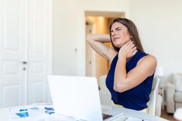 Фото Молодая деловая женщина, страдающая от боли в шее, чувствует усталость и стресс разочарованная молодая женщина, страдающая от боли в шее, массирует шею, сидя на рабочем месте в домашнем офисе
