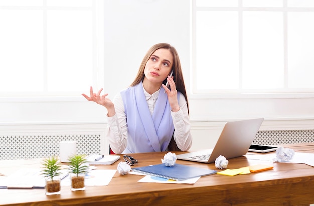 Young businesswoman talking by phone with papers, sitting at modern office workplace. Business consulting.