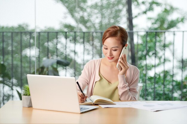 Young businesswoman talking on phone while using laptop at table in office