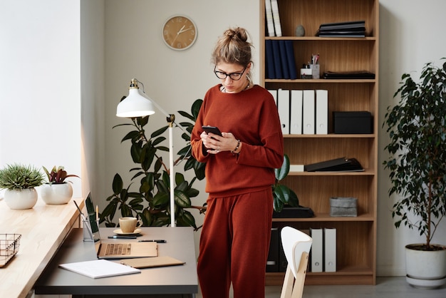 Young businesswoman texting message on mobile phone while standing near her workplace with laptop during her work at office