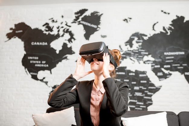 Photo young businesswoman trying virtual reality glasses sitting indoors with world map on the background